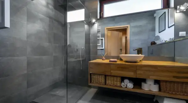 Salle de bain moderne avec douche à l'italienne, carrelage gris, meuble en bois, vasque et miroir. L'étanchéité de la douche italienne est assurée par un receveur discret.