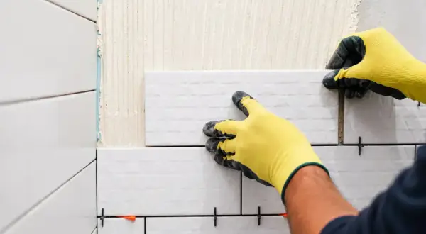 Mains gantées jaunes en pleine pose carrelage mural blanc. Des croisillons maintiennent l'espacement.