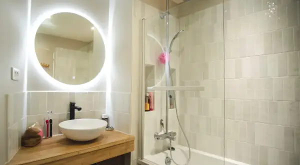 Salle de bain sans fenêtre, lumineuse avec un grand miroir rond éclairé, une vasque moderne et une douche à l'italienne.