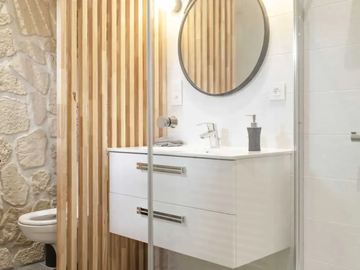 Salle de bain moderne avec un lavabo suspendu, un miroir rond et une **installation WC** discrète derrière un paravent en bois.