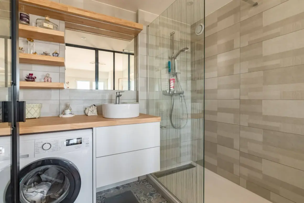 Coin buanderie dans salle de bain moderne avec lave-linge, lavabo sur meuble blanc et douche vitrée.