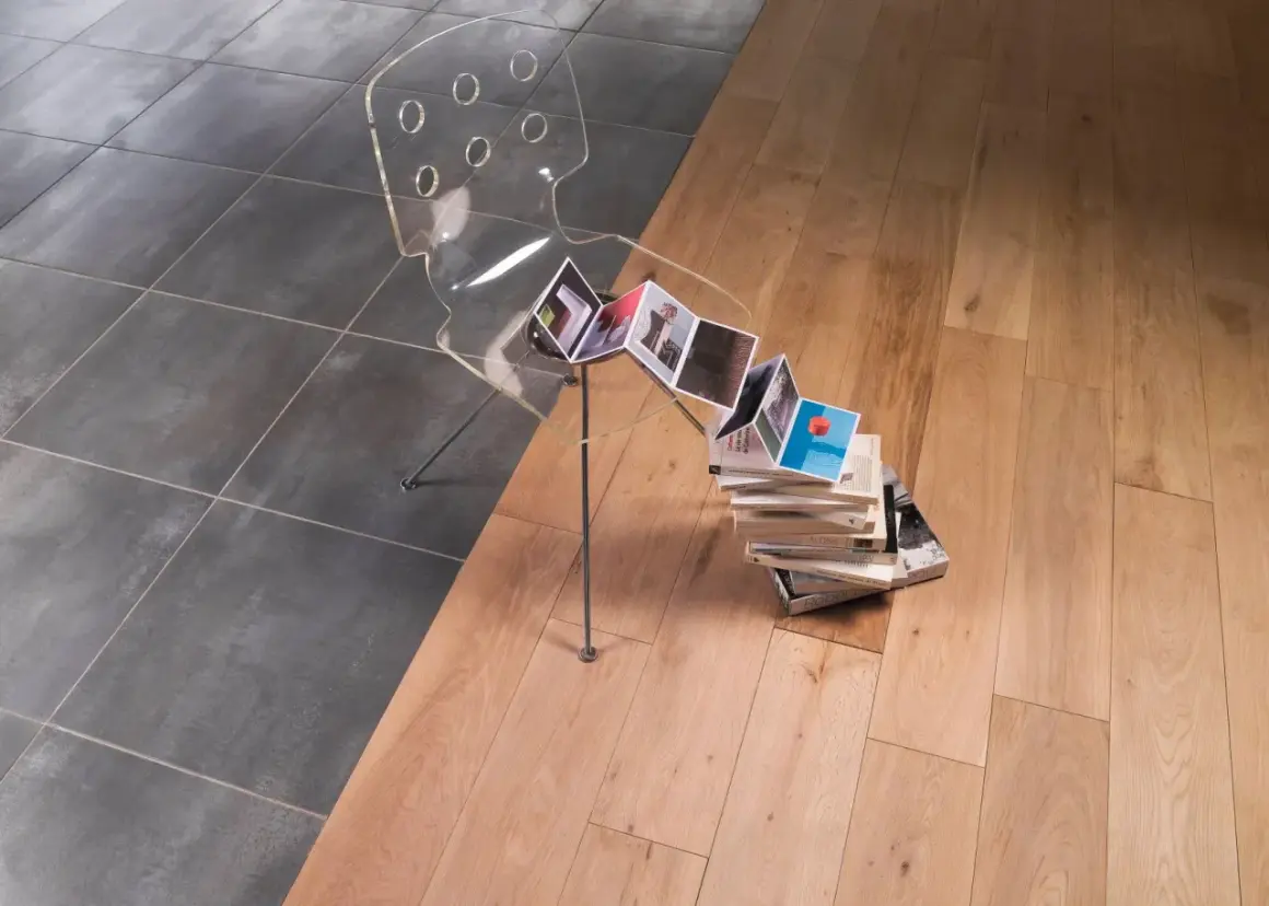 Une barre de seuil discrète sépare un carrelage sombre d'un parquet clair. Une chaise transparente et des livres créent une scène artistique.