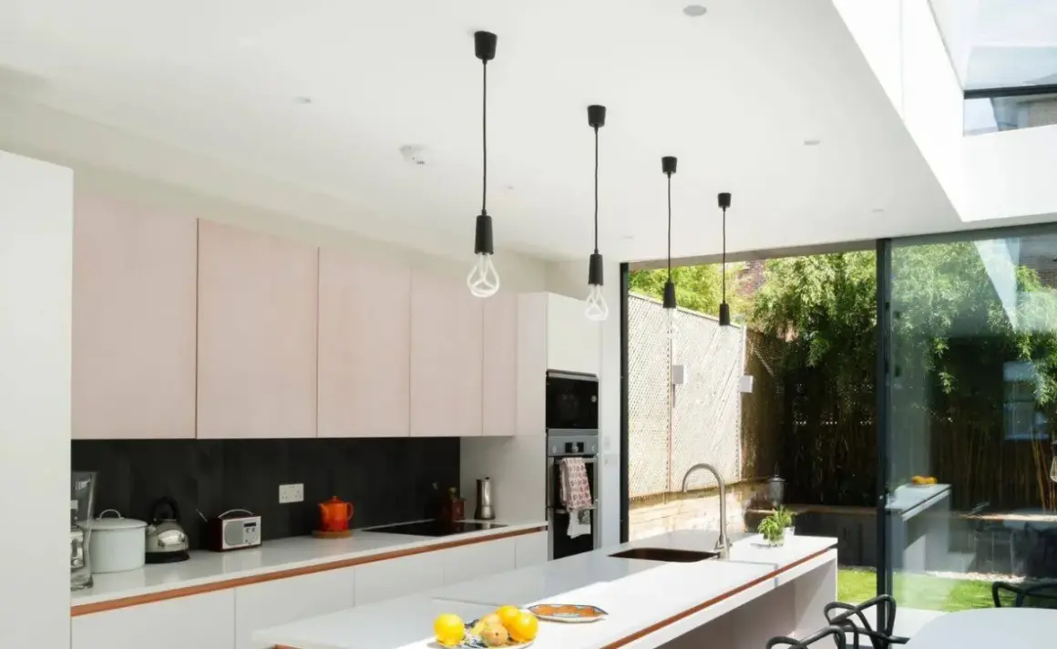 Cinq suspensions noires au design épuré, quel luminaire pour cuisine moderne ! Elles éclairent une cuisine lumineuse avec ses meubles roses et sa grande baie vitrée.