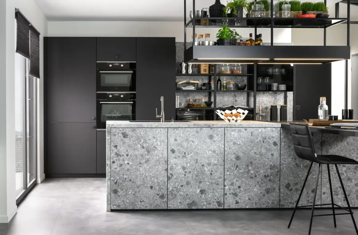 Cuisine moderne aux façades grises et à l'îlot central en sol terrazzo. Électroménagers intégrés et étagères noires remplies d'objets décoratifs.