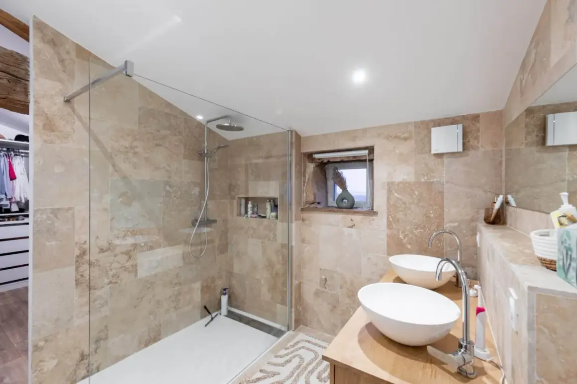 Salle de bain moderne avec douche à l'italienne, double vasque et carrelage en travertin. Une transformation réussie de rénovation salle de bain avant/après.