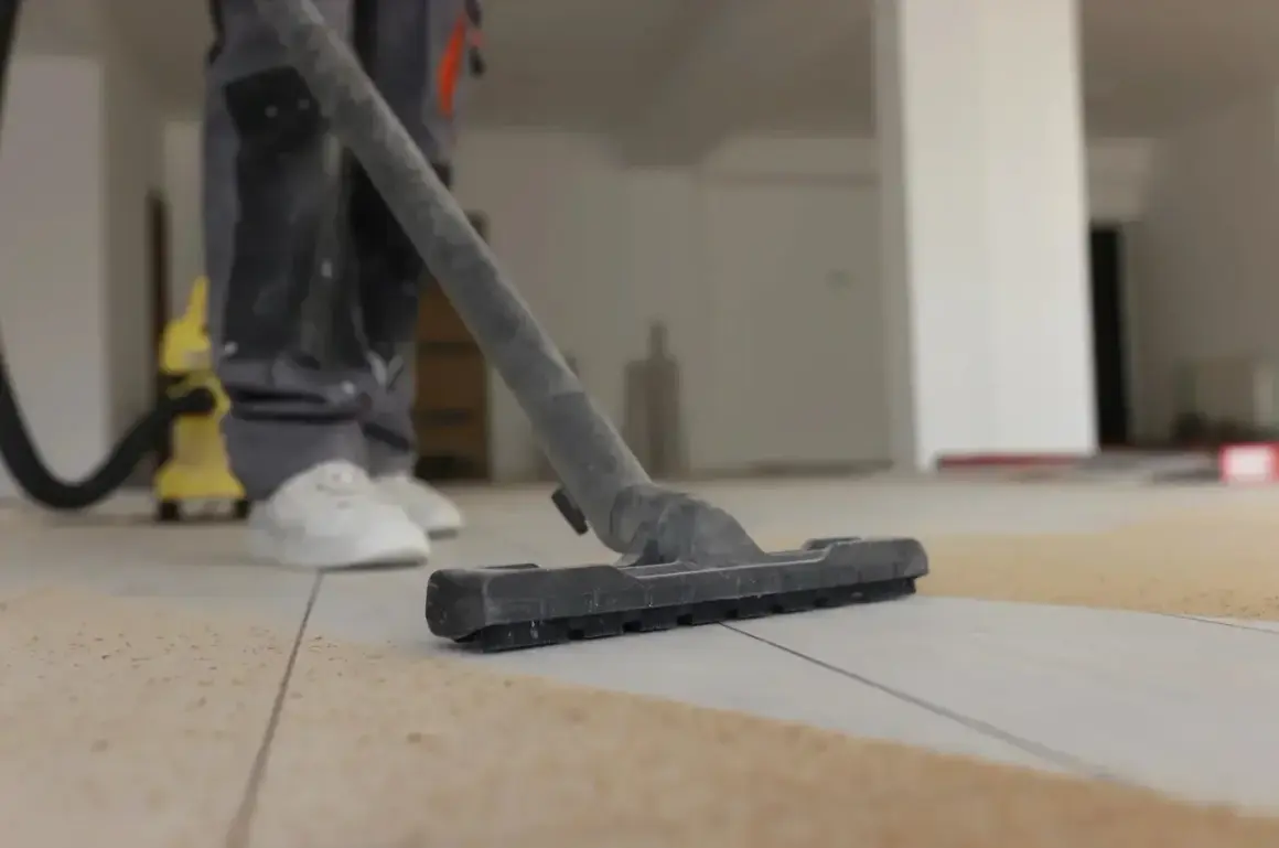 Un ouvrier utilise un aspirateur pour nettoyer le carrelage après le jointoiement. La poussière et les débris sont aspirés.