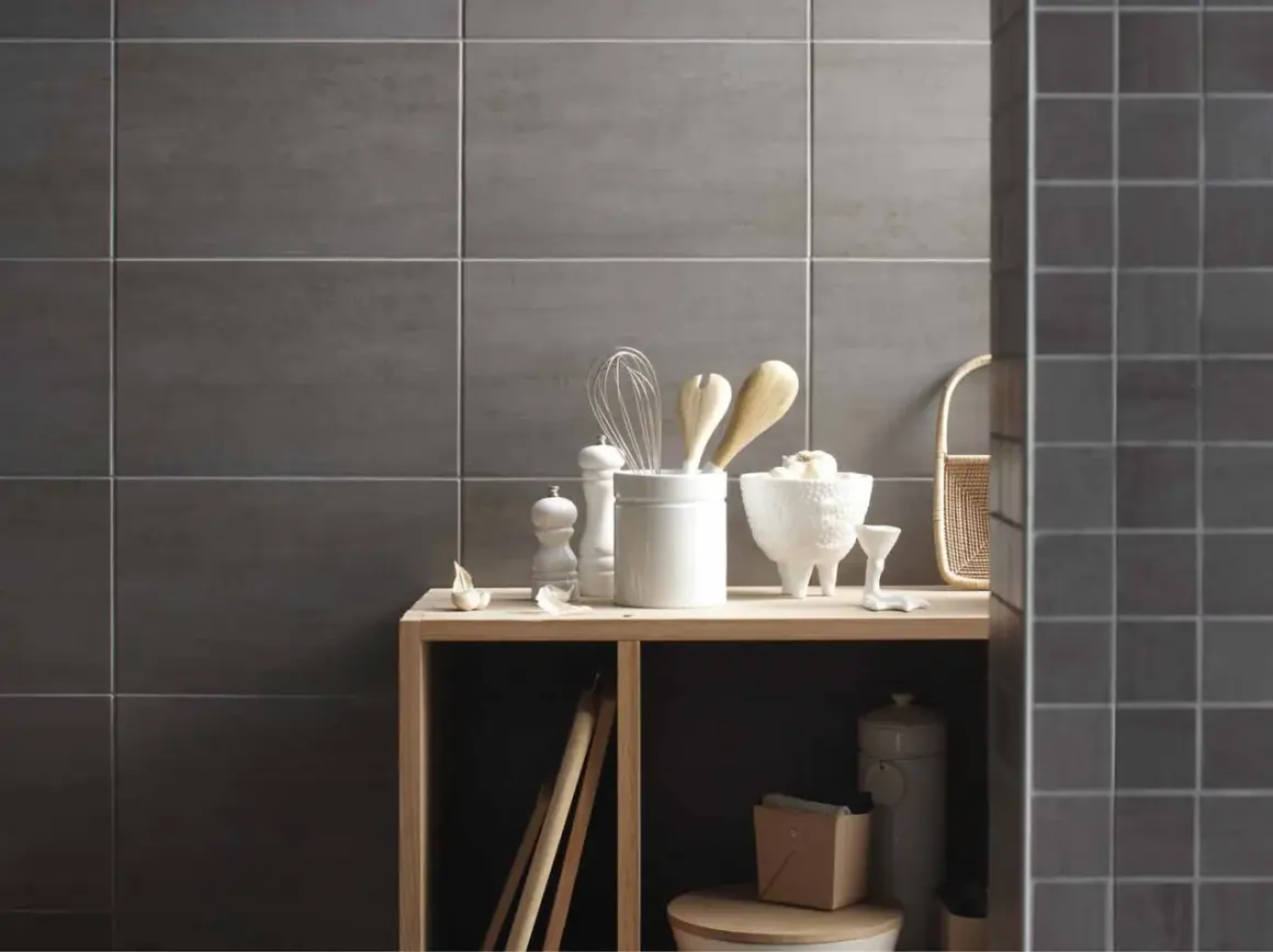 Sur un mur en carrelage gris, des ustensiles de cuisine et des objets décoratifs sont disposés sur une étagère en bois. La faïence du mur de droite est plus petite.