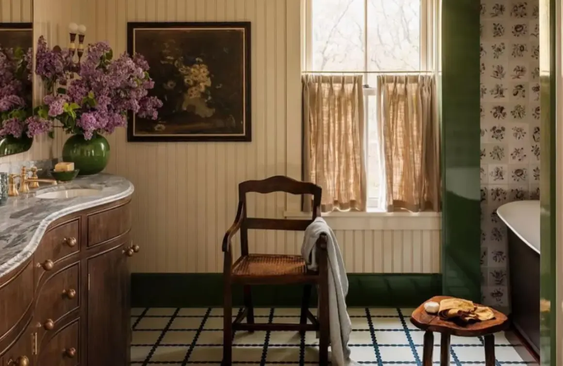 Ambiance de salle de bain rustique avec un lavabo en marbre, des lilas violets, un tableau ancien et une chaise en bois.