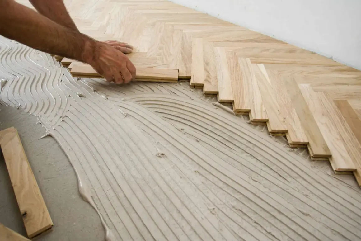 Mains d'un artisan en train de poser du parquet en chevrons sur un lit de colle ondulée.