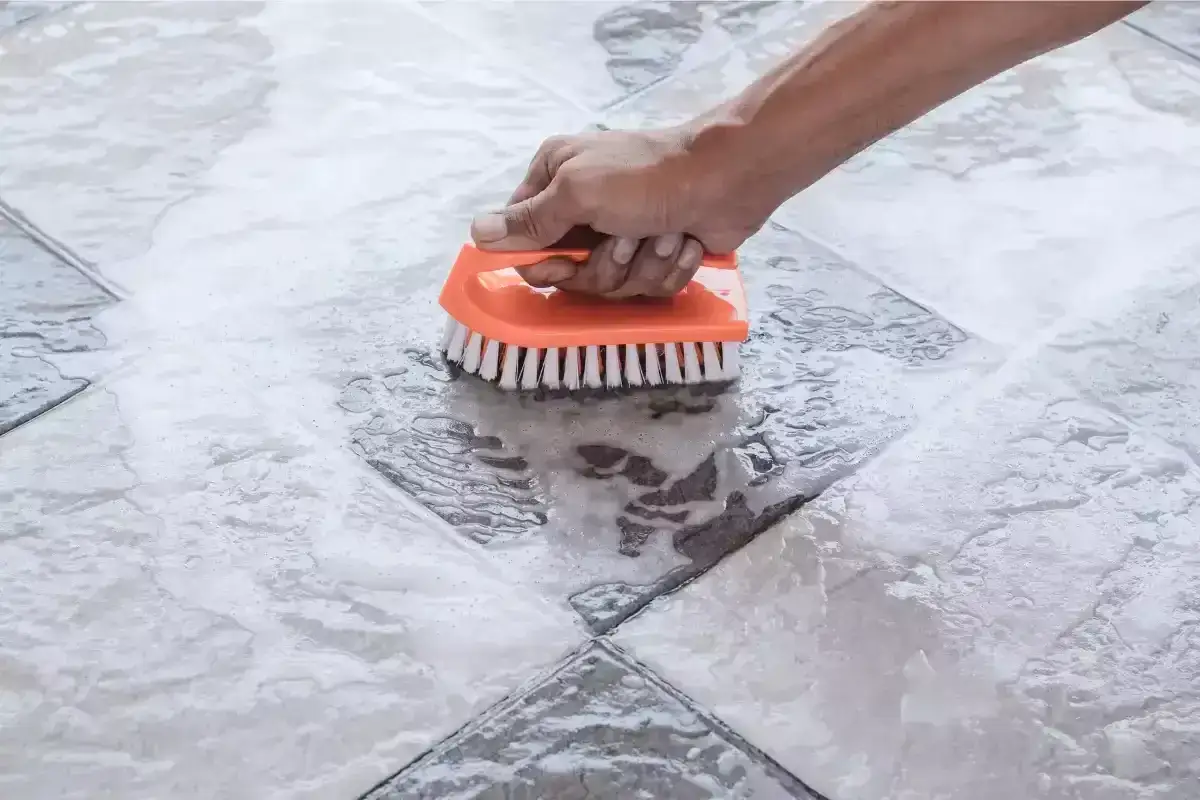 Main tenant frotte une tache acide sur carrelage avec une brosse orange.