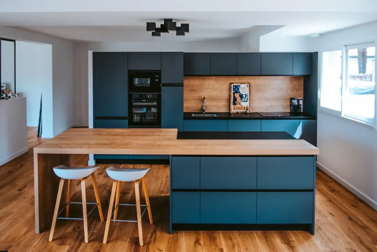 Cuisine ouverte en L, moderne et épurée, avec des meubles bleu pétrole, un îlot en bois clair et deux tabourets.