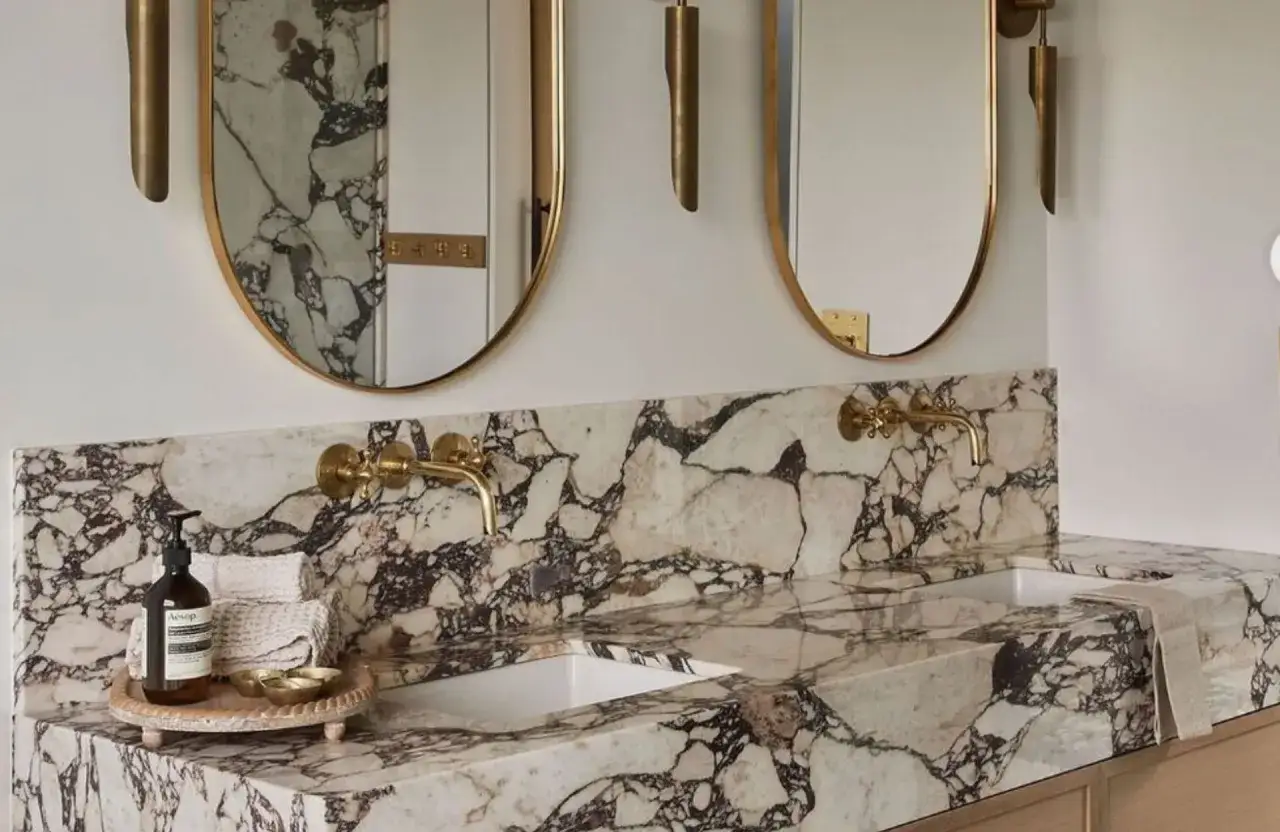 Une salle de bain luxueuse avec un plan de travail et un dosseret en marbre veiné. L'idée salle de bain marbre est sublimée par des robinets dorés et des miroirs ovales.