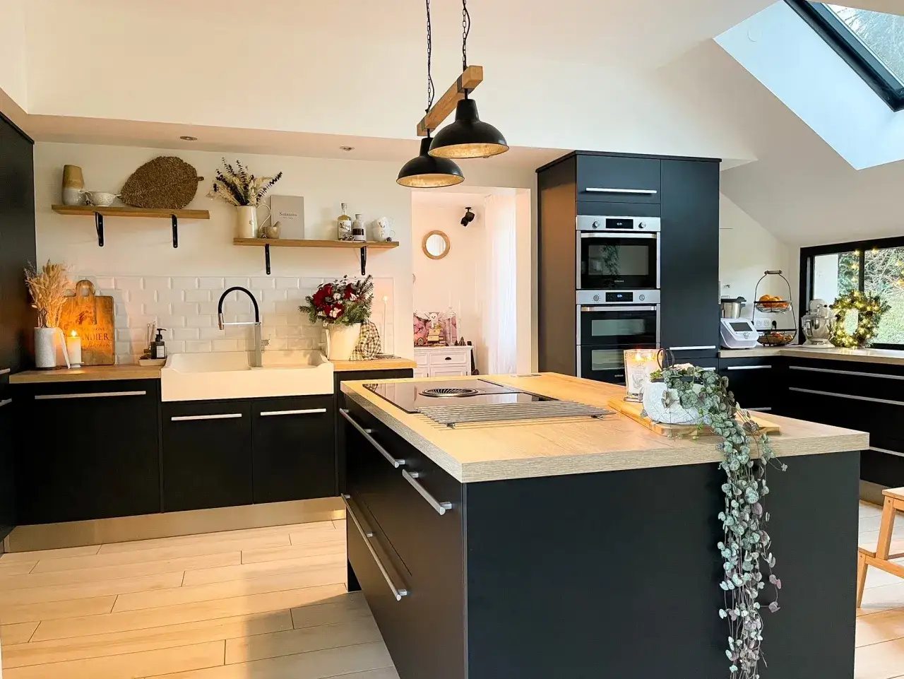Cuisine moderne avec îlot central en bois clair, meubles noirs, évier blanc et luminaires industriels. Une belle idée aménagement cuisine.