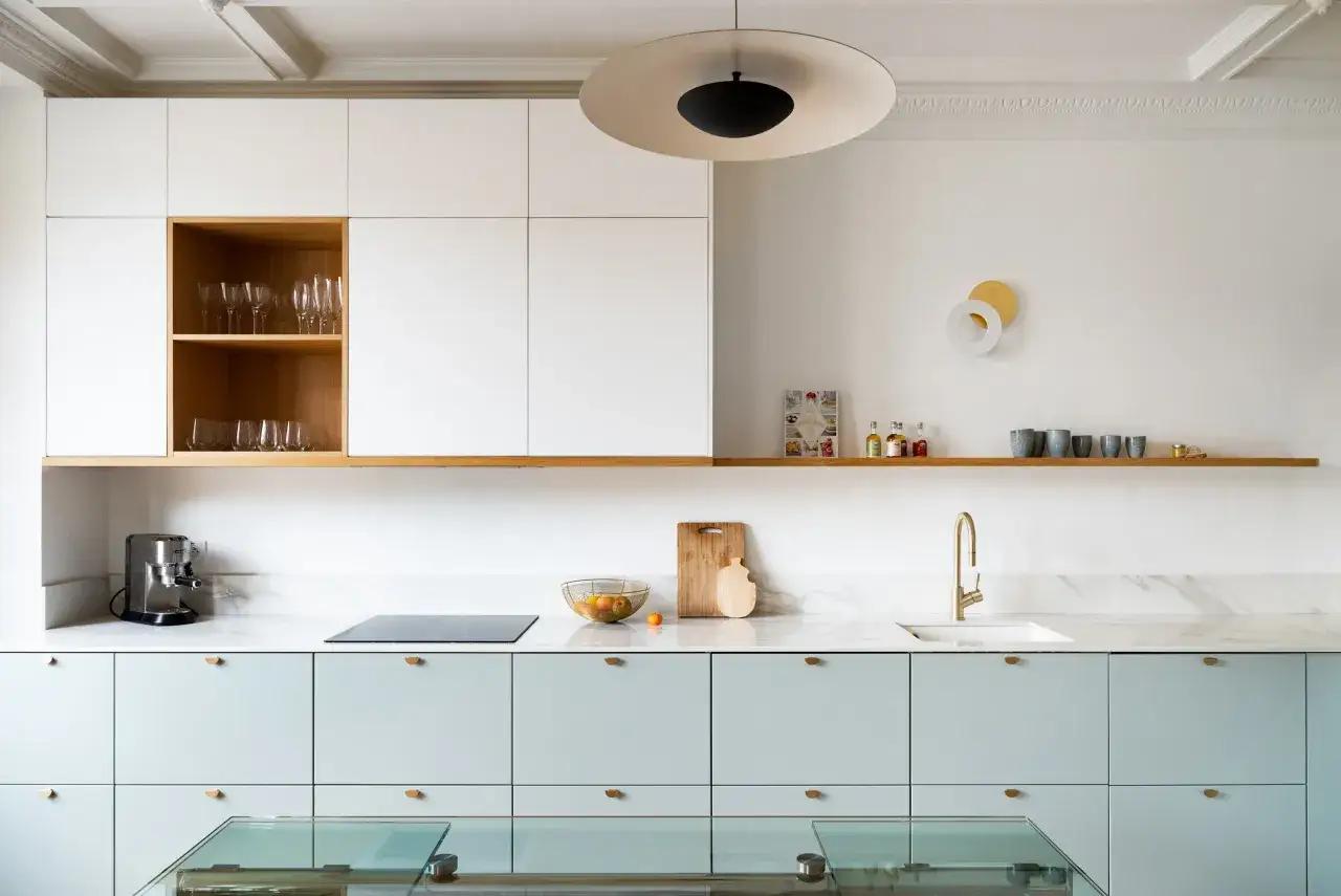 Une cuisine moderne aux façades bleu pastel et aux accents de bois. Des verres sont rangés dans une niche en bois, tandis qu'un plan de travail en marbre accueille une cafetière et un évier doré.