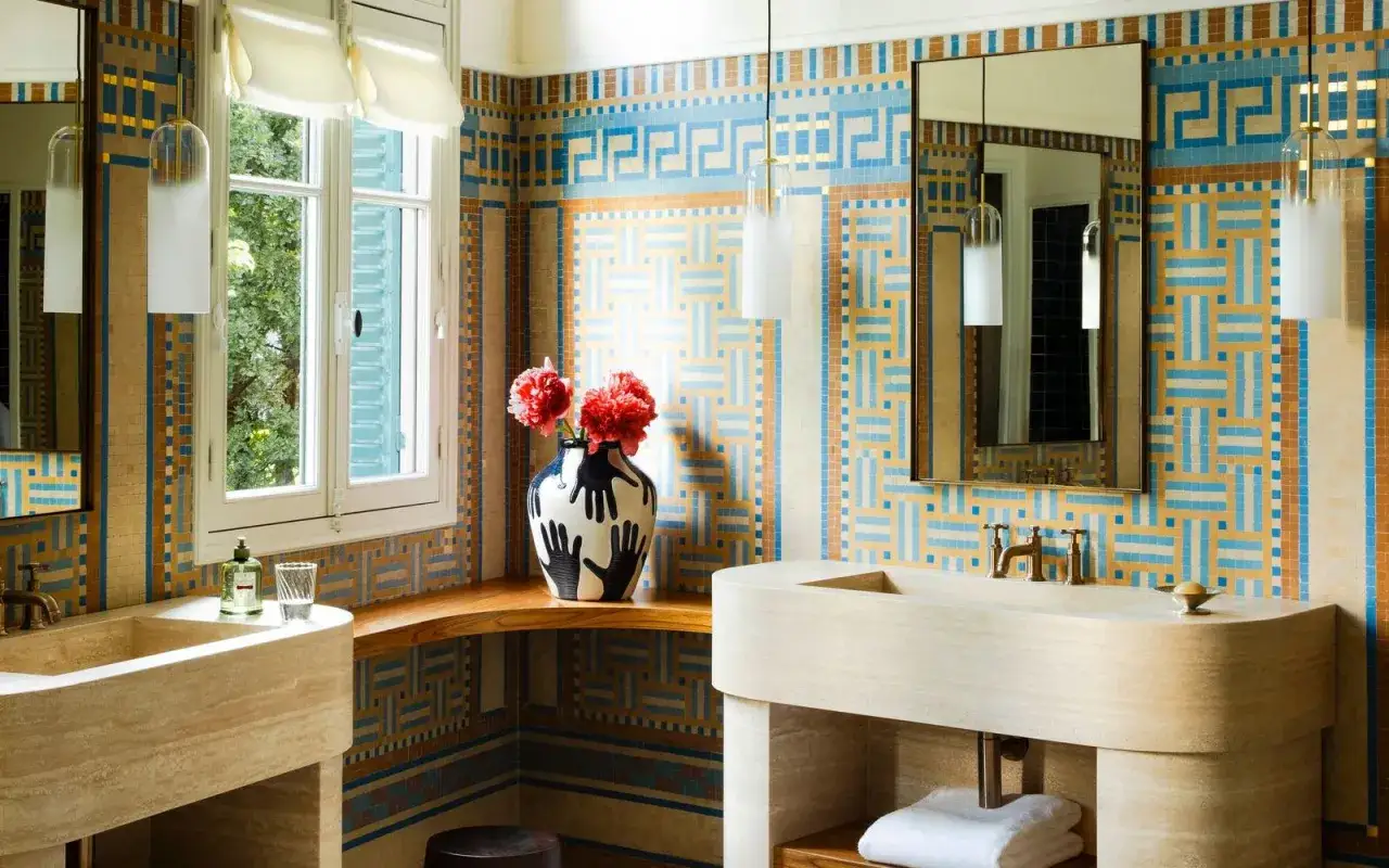 Élégante salle de bain art déco aux murs carrelés de motifs géométriques bleus et ocre. Lavabos en pierre, robinetterie dorée et vase décoratif.