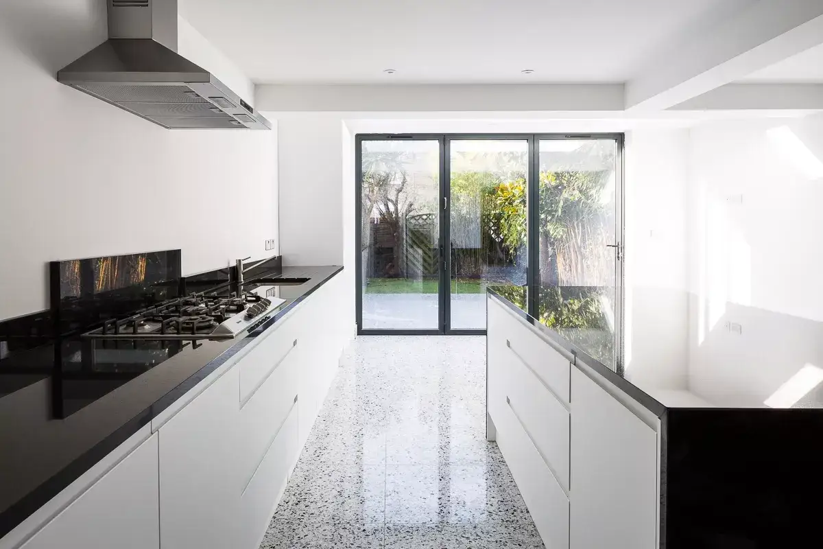 Cuisine moderne avec un sol en terrazzo étincelant, des plans de travail noirs et des armoires blanches épurées. Des portes vitrées s'ouvrent sur un jardin verdoyant.