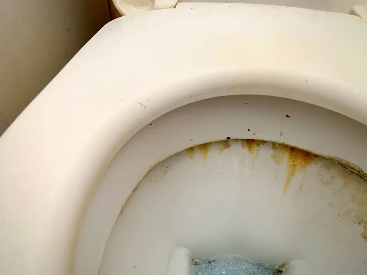 Traces de calcaire brunâtres et jaunâtres sur le rebord intérieur d'une toilette blanche.