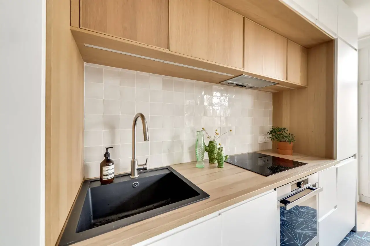 Cuisine moderne avec un évier noir, une plaque de cuisson et des armoires en bois clair. Le mur est recouvert d'une faience blanche texturée.