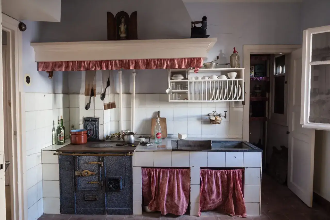 Cuisine am&eacute;nag&eacute;e avec des vieux meubles, un po&ecirc;le ancien, une vaisselle en bois et des rideaux &agrave; carreaux rouges.