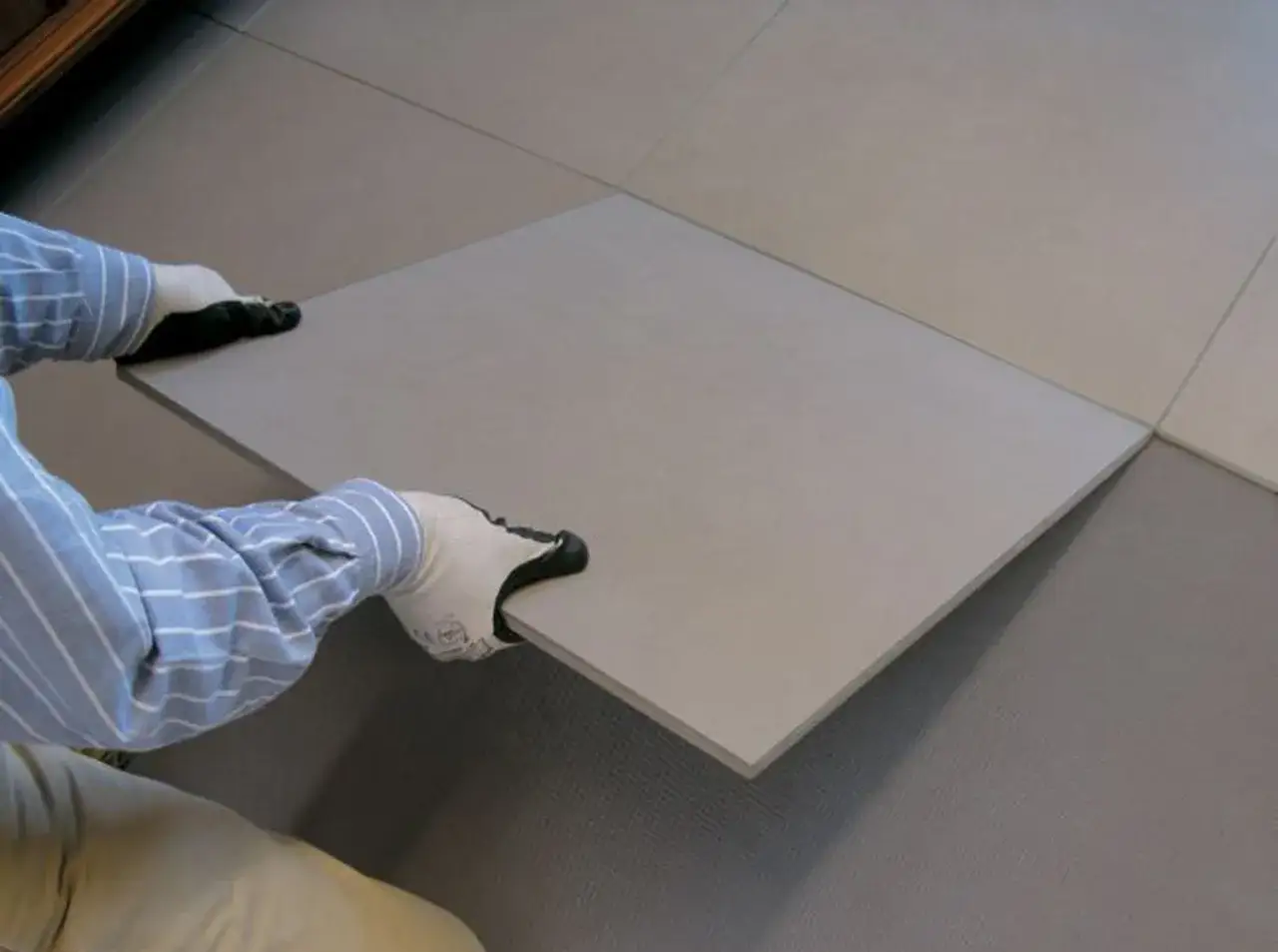 Pose d'une nouvelle dalle de carrelage sur carrelage existant. Mains gantées tenant la nouvelle pièce.