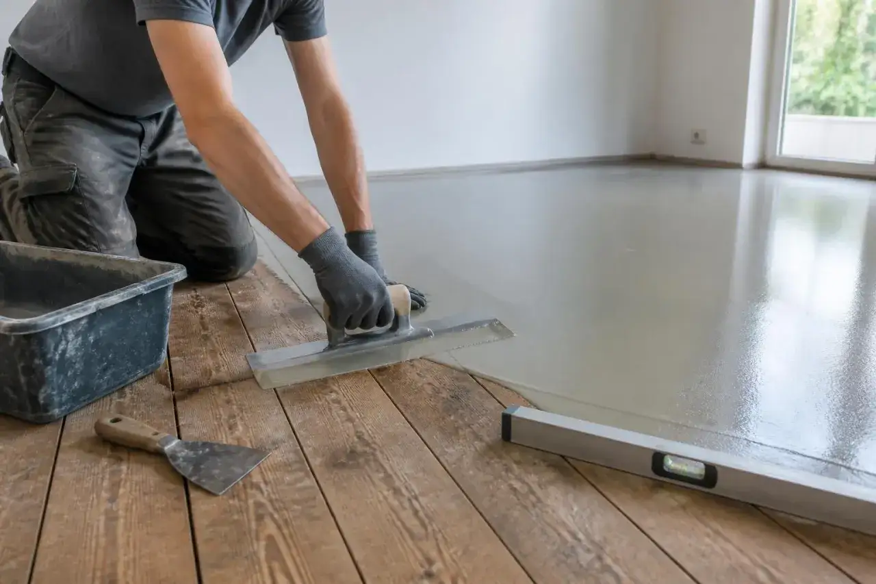 Un artisan réalise un ragréage sur plancher bois, lissant la chape avec une taloche pour une surface parfaite.