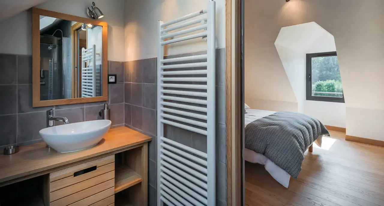 Salle de bain sous pente avec douche &agrave; faible hauteur, lavabo moderne et radiateur blanc. Chambre attenante avec lit douillet.
