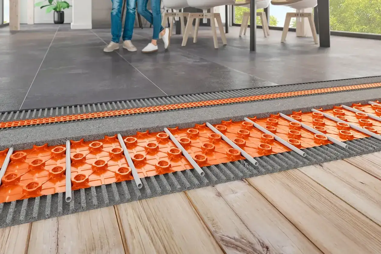 Vue en coupe d'un syst&egrave;me de chauffage par le sol &eacute;lectrique, montrant les tubes orange int&eacute;gr&eacute;s dans une couche isolante sous un rev&ecirc;tement de sol moderne.