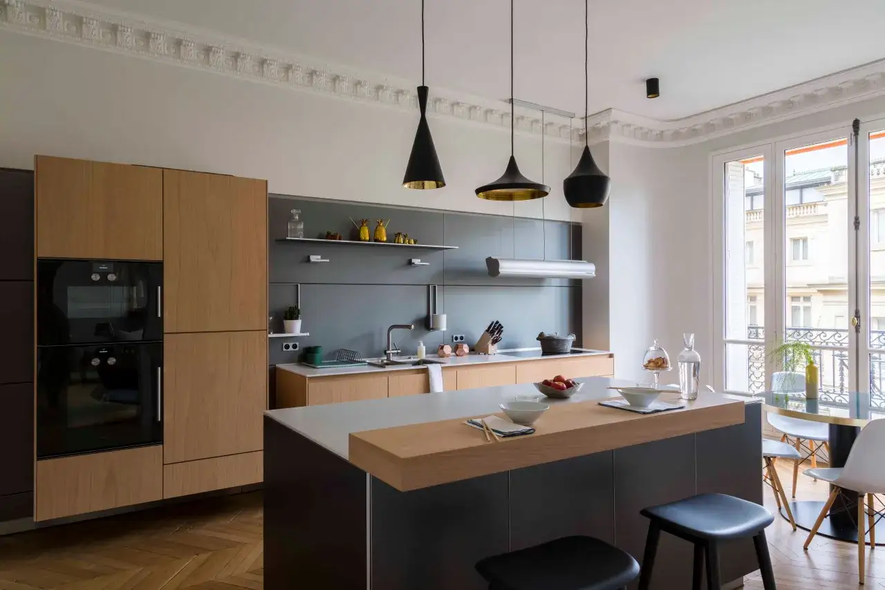 Cuisine moderne avec îlot central, placards en bois clair et carrelage marron foncé au sol. Luminaires noirs suspendus.