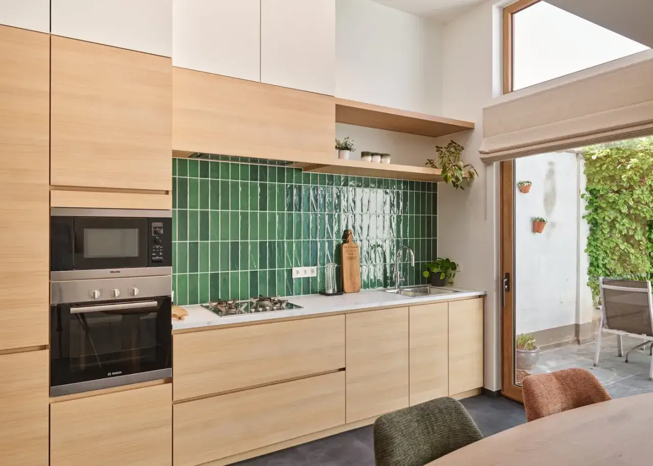 Cuisine moderne avec crédence verte brillante et meubles en bois clair. Un four et un micro-ondes intégrés. La cuisine en parallèle s'ouvre sur un patio verdoyant.