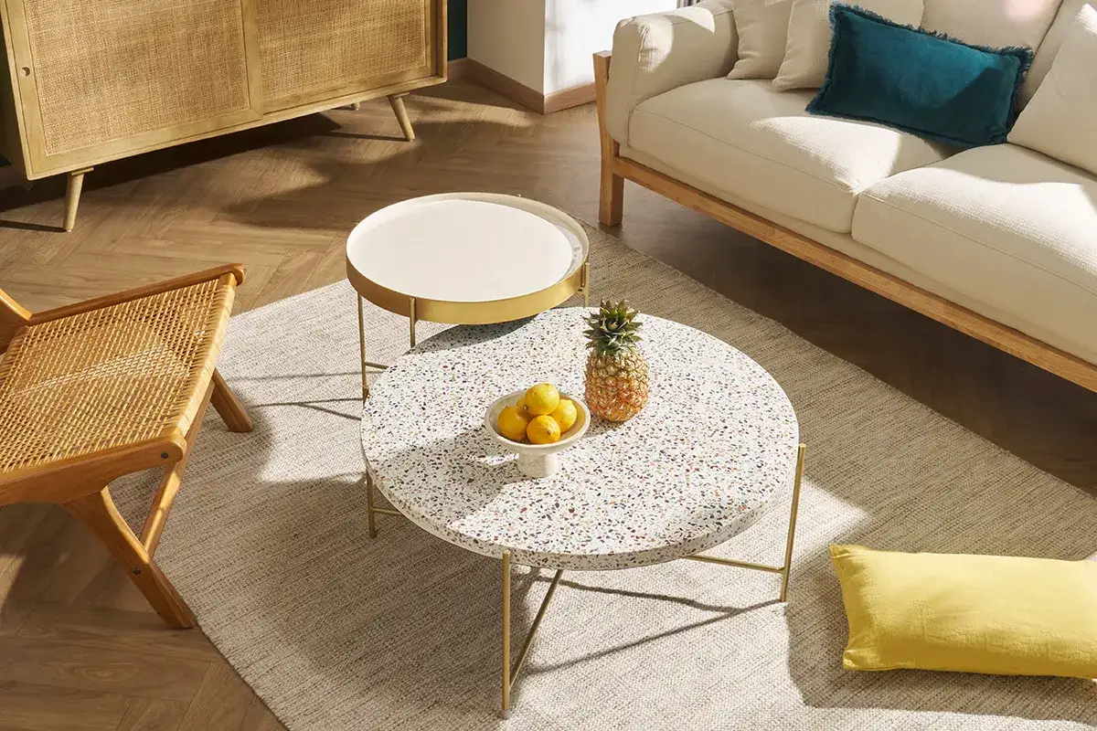Un salon cosy avec un canapé clair, un fauteuil en rotin et une table basse au plateau en terrazzo coloré, ornée d'une ananas et de citrons.