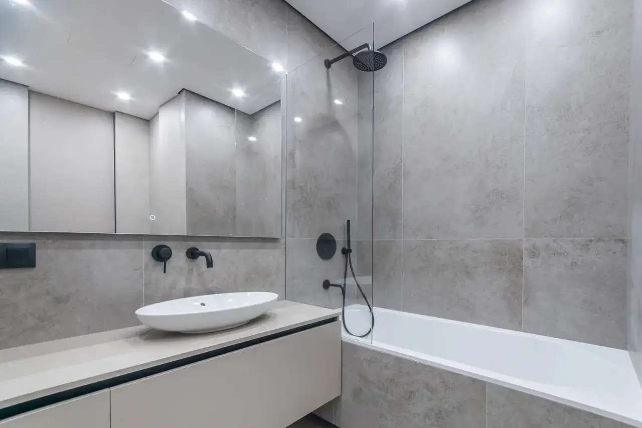 Salle de bain moderne avec lavabo ovale, robinetterie noire et baignoire. Le carrelage autour de la baignoire est gris et texturé.