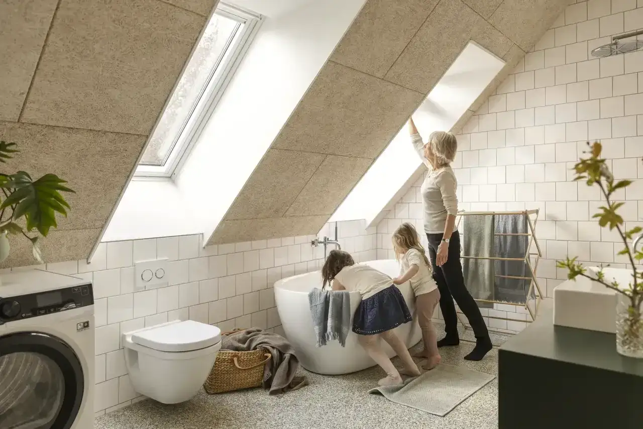 Salle de bain sous pente avec wc suspendu. Une femme et deux enfants admirent la lumière entrant par la fenêtre de toit.