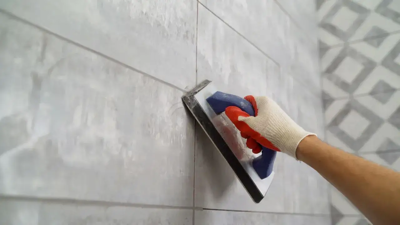 Main d'un carreleur appliquant du joint carrelage gris sur un mur de carreaux gris.