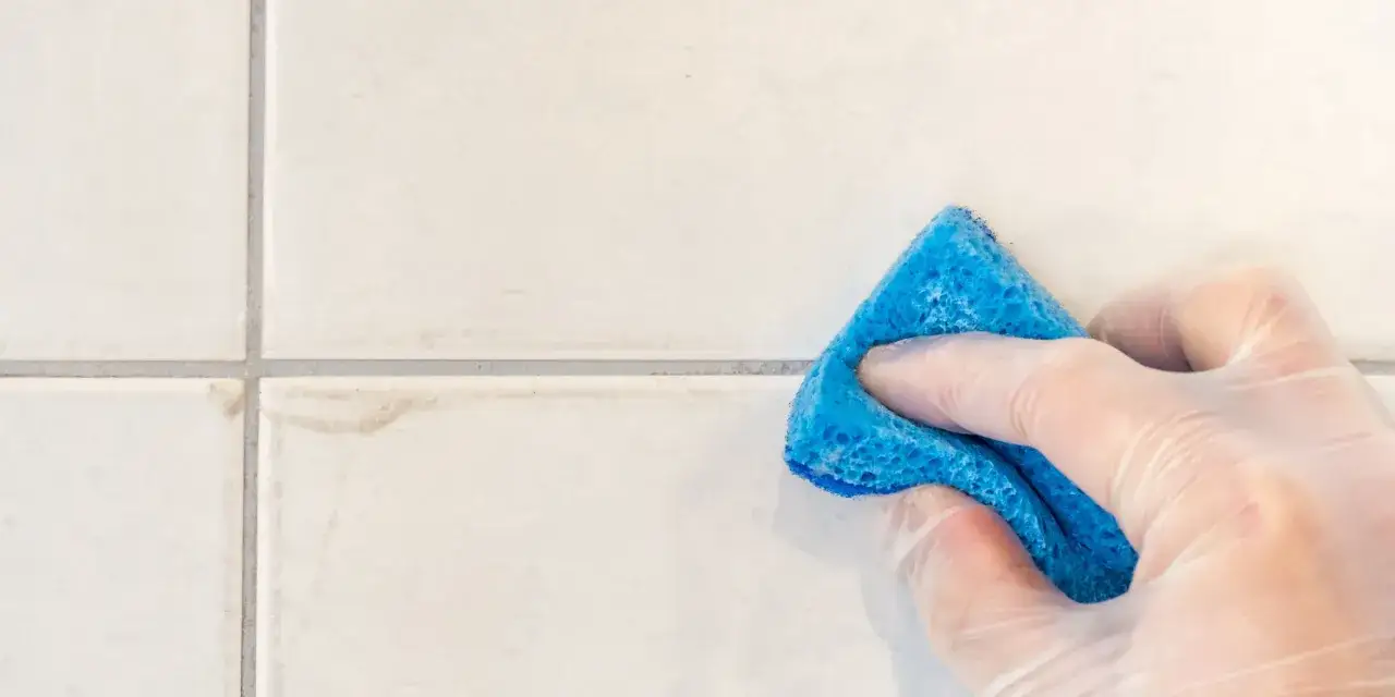 Main tenant utilise une éponge bleue pour nettoyer du javel sur carrelage, éliminant les taches tenaces.