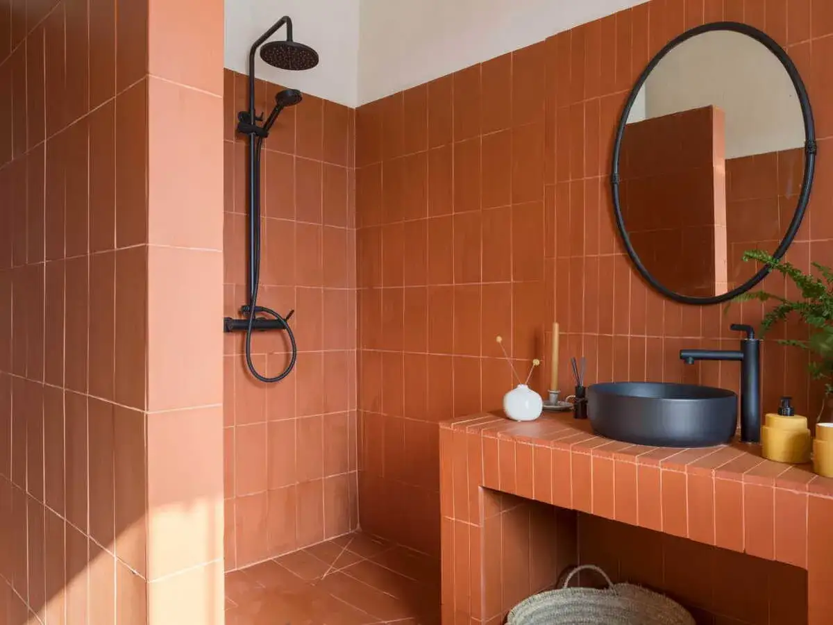 Salle de bain tomette aux tons chauds. Douche noire, vasque moderne et miroir rond sur fond de carrelage ocre.