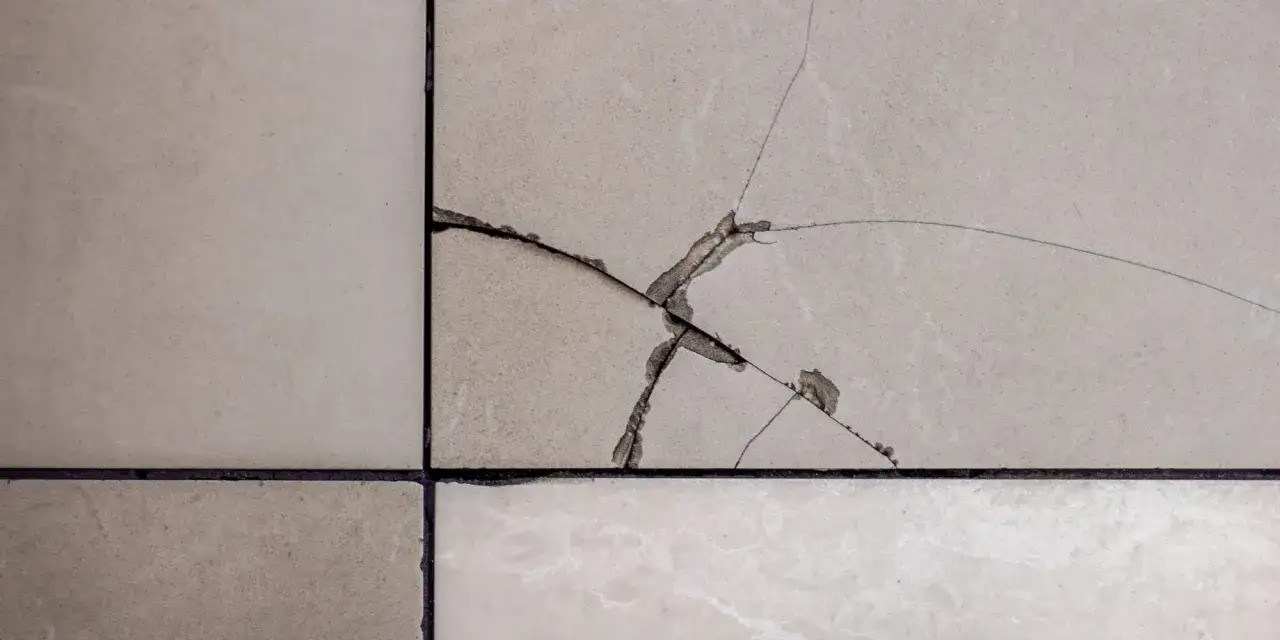 Gros plan sur une fissure carrelage beige, révélant des éclats et des lignes fines traversant la surface.