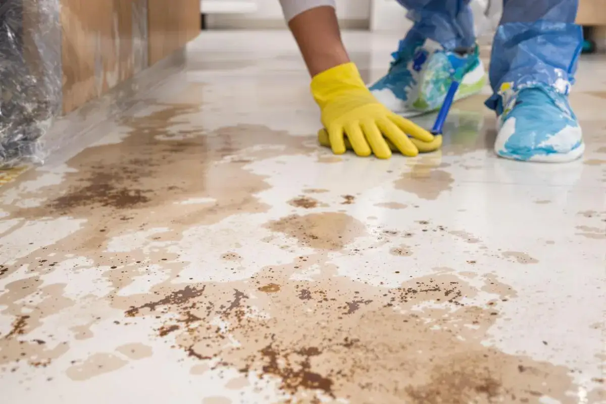 Nettoyage d'un déversement sur du carrelage. Une main gantée de jaune utilise un outil pour nettoyer des taches brunes, peut-être dues à de l'acide chlorhydrique.