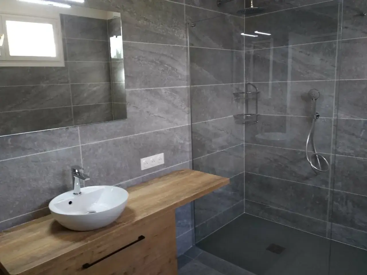 Salle de bain gris clair et gris foncé. Douche moderne avec paroi en verre, lavabo sur meuble en bois, et miroir.