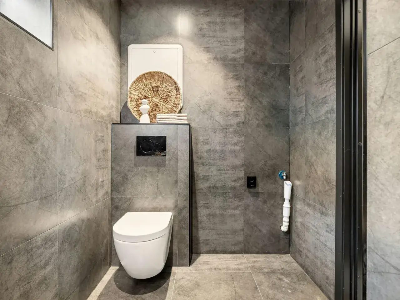 Installation WC moderne avec cuvette suspendue et plaque de commande noire. Le mur est carrelé de grands carreaux gris effet béton.