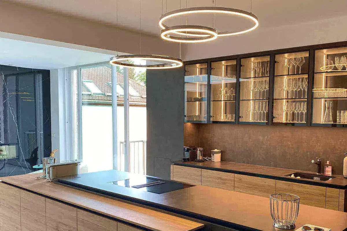 Quel luminaire pour cuisine ! Deux suspensions circulaires modernes éclairent cette cuisine design avec îlot central et placards vitrés remplis de verres.