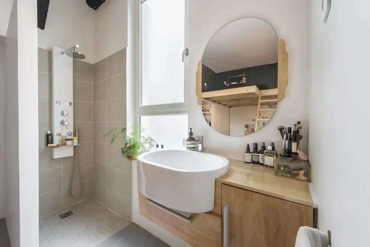Salle de bain blanc et bois, avec douche moderne, vasque design et miroir rond reflétant un lit mezzanine.