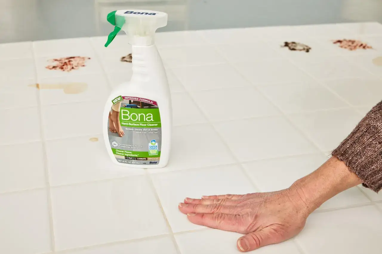 Une main efface des taches sur un carrelage blanc, à côté d'un flacon Bona. Obtenez un carrelage brillant sans trace avec ce nettoyant.