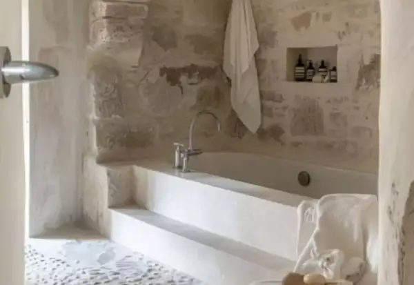Bain relaxant dans une **salle de bain méditerranéenne** aux murs de pierre brute. Serviette blanche suspendue, robinetterie moderne.