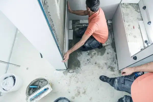 Un ouvrier réalise l'installation d'une douche, posant des panneaux muraux. Outils et matériaux de construction sont visibles au sol.