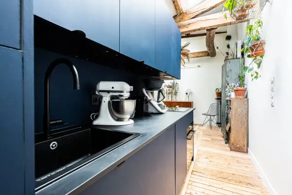 Cuisine moderne avec armoires bleues, évier noir et robots culinaires. L'espace offre une entrée directe dans la cuisine, avec des poutres apparentes et des plantes vertes.