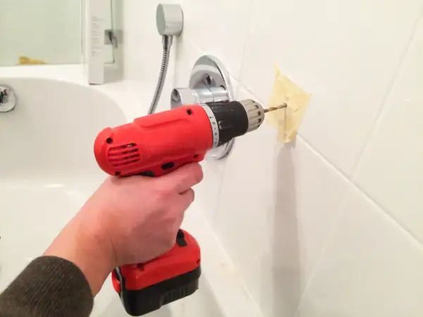 Une main tient une perceuse rouge pour percer du carrelage blanc, avec du ruban adhésif pour guider le foret.