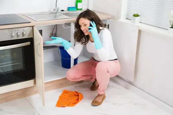 Une femme en gants bleus, au téléphone, tente de colmater une fuite d'eau sous son évier, un seau bleu à ses pieds.
