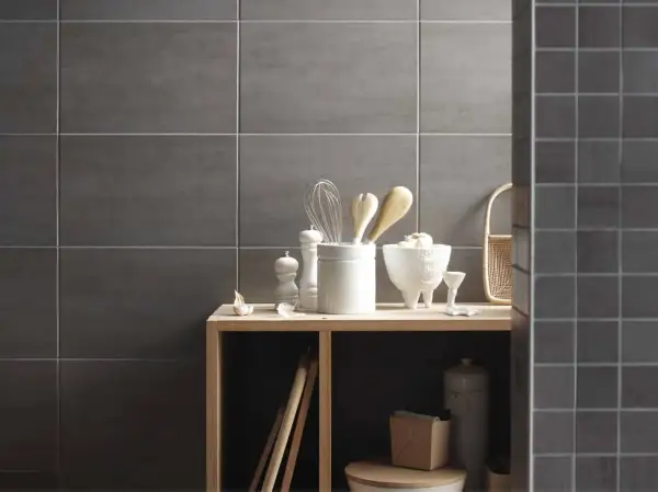 Sur un mur en carrelage gris, des ustensiles de cuisine et des objets décoratifs sont disposés sur une étagère en bois. La faïence du mur de droite est plus petite.