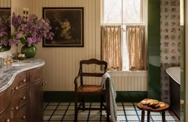 Ambiance de salle de bain rustique avec un lavabo en marbre, des lilas violets, un tableau ancien et une chaise en bois.