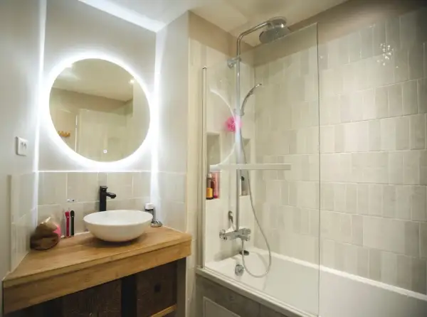 Salle de bain sans fenêtre, lumineuse avec un grand miroir rond éclairé, une vasque moderne et une douche à l'italienne.